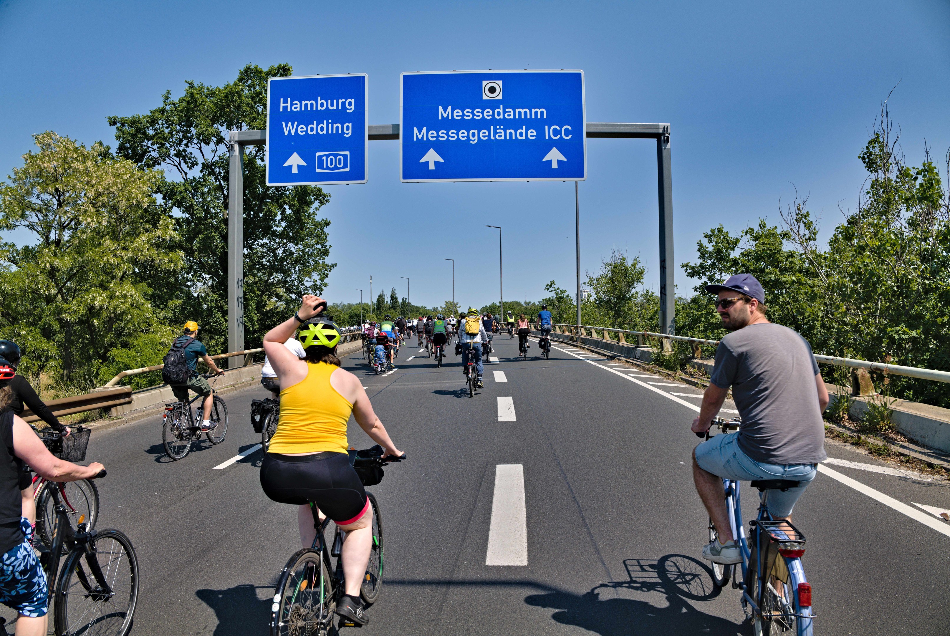 Eine Gruppe von Radfahrern mit Helmen fährt auf einer Straße mit einer Begrenzung auf einer Seite und Bäumen auf der anderen, unter einem klaren blauen Himmel mit Laternen im Hintergrund; ein Schild oben zeigt eine Radtour in Hamburg.