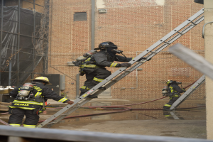 Feuerwehrleute in Helmen und Ausrüstung klettern an einer Leiter vor einem Backsteingebäude mit Rohren auf dem Boden und einer Metallstange unten, mit einem anderen Gebäude mit Fenstern und einem Netz im Hintergrund.