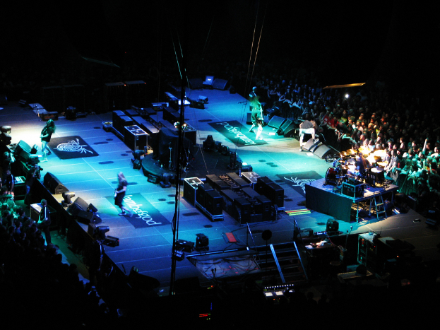 Eine Gruppe von Menschen, die auf einer Bühne mit Lautsprechern, Scheinwerfern und Mikrofonen performs, umgeben von einer großen Menge im dunklen Hintergrund.