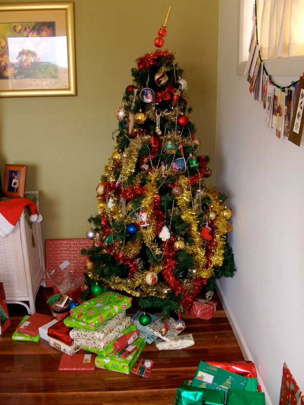 Ein geschmückter Weihnachtsbaum mit Geschenken am Fuße, ein Photo-Rahmen an der linken Wand und ein Vorhang am rechten Fenster.