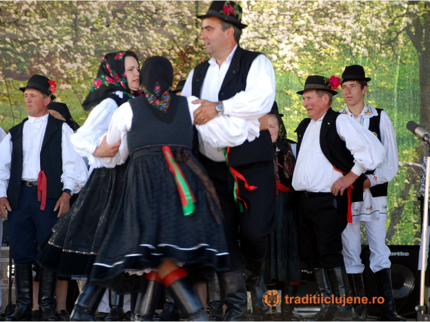 Eine Gruppe von Menschen in traditioneller ungarischer Kleidung tanzt auf einer Bühne mit Mikrofonen, Lautsprechern und Bäumen im Hintergrund, und Text am unteren Rand des Bildes.