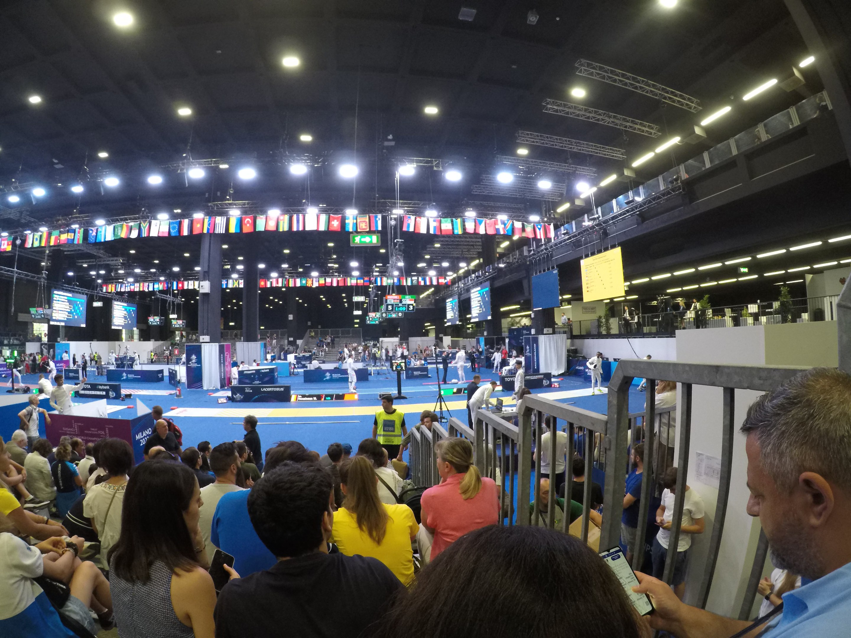 Eine große Menschenmenge sitzt und steht in einer Indoor-Sportarena mit Bildschirmen, Fahnen und Deckenbeleuchtung im Hintergrund.
