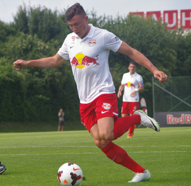 Ein Mann in einem Fussballtrikot schießt einen Ball auf einem grünen Feld mit Bäumen und einem klaren blauen Himmel im Hintergrund, wobei ein Schild mit der Aufschrift "RB Leipzig" sichtbar ist.