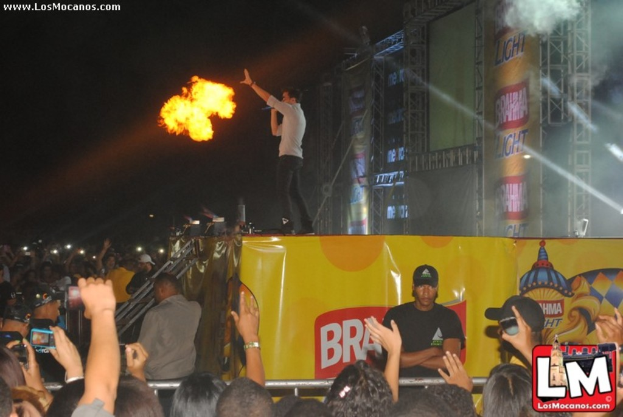 Eine Gruppe von Menschen, einige mit Kameras, mit einem Mann in der Mitte, der einem Feuer gegenübersteht, umgeben von Metallstangen, Absperrungen und Rauch, mit einem Wasserzeichen oben links und einem Logo unten rechts.