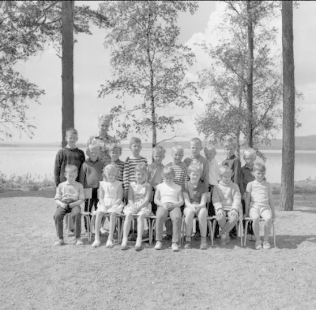 Schwarz-weißes Foto einer Gruppe von Kindern, die auf Stühlen sitzen und vor einem See stehen, mit Bäumen, Pflanzen, Hügeln und Wolken im Hintergrund, beschriftet mit "1940er Kinder am See."