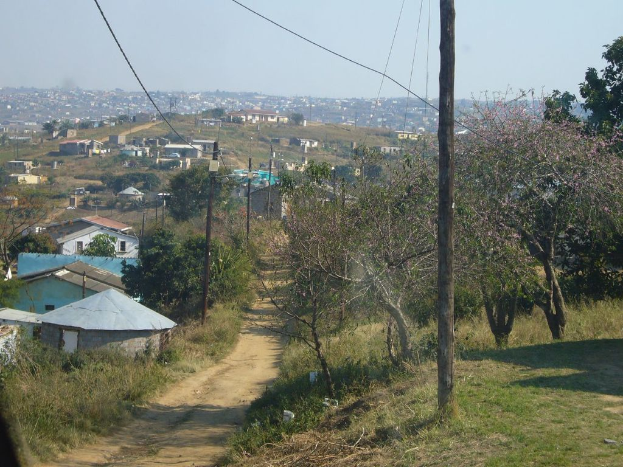 Ein Weg mit Grün und Bäumen auf beiden Seiten, mit Pfählen und Gebäuden im Hintergrund.