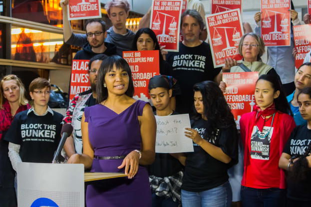 Eine Black Lives Matter-Aktivistin spricht vor einem Publikum mit Mikrofon an einem Rednerpult, während das Publikum Schilder hält.