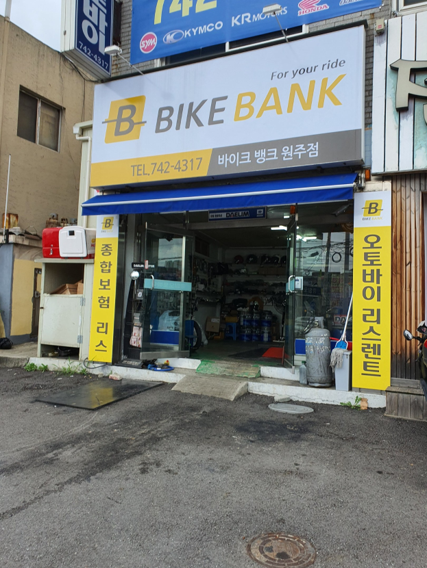 Außenansicht eines Fahrradgeschäfts namens "Bike Bank" mit parkenden Fahrzeugen davor, einer Straße rechts daneben und Schautafeln drinnen.