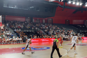 Eine Gruppe von Menschen, die Basketball in einer Turnhalle spielen, mit einem Basketballkorb auf der linken Seite und Zuschauern, die auf Stühlen sitzen, im Hintergrund.
