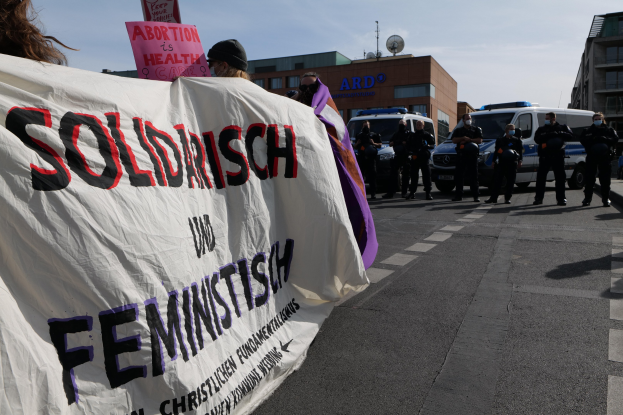 Gruppe von Menschen marschiert durch eine Straße und hält ein 'Solidarität und Feminismus' -Schild, mit parkenden Fahrzeugen und Gebäuden im Hintergrund bei klarem blauem Himmel.