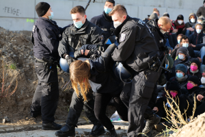 Gruppe maskierter Personen in Mützen, einige bewaffnet, umgeben eine Frau, die von der Polizei eskortiert wird, mit Pflanzen, Steinen und einer Wand mit Protesttext im Hintergrund.