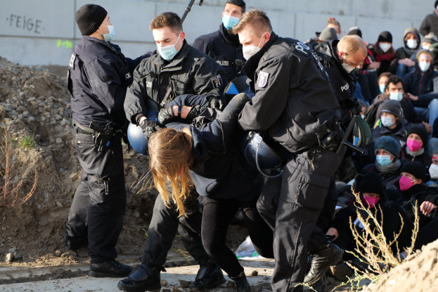 Gruppe maskierter Personen in Mützen, einige bewaffnet, umgeben eine Frau, die von der Polizei eskortiert wird, mit Pflanzen, Steinen und einer Wand mit Protesttext im Hintergrund.