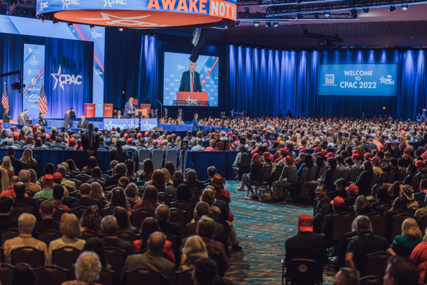 Eine große Menschenmenge auf Stühlen sitzend, die einer Bühne gegenüberstehen, mit einigen Menschen auf dem Podium und auf der linken Seite, blaue Vorhänge und ein Bildschirm im Hintergrund und eine orangefarbene Tafel oben, die das CPAC 2020-Event am 25. Oktober anzeigt.