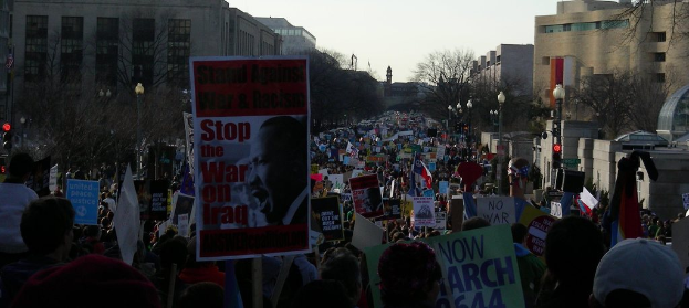 Menschen demonstrieren auf einer Straße mit Plakaten, mit Gebäuden, Bäumen, Laternenmasten und dem Himmel im Hintergrund.