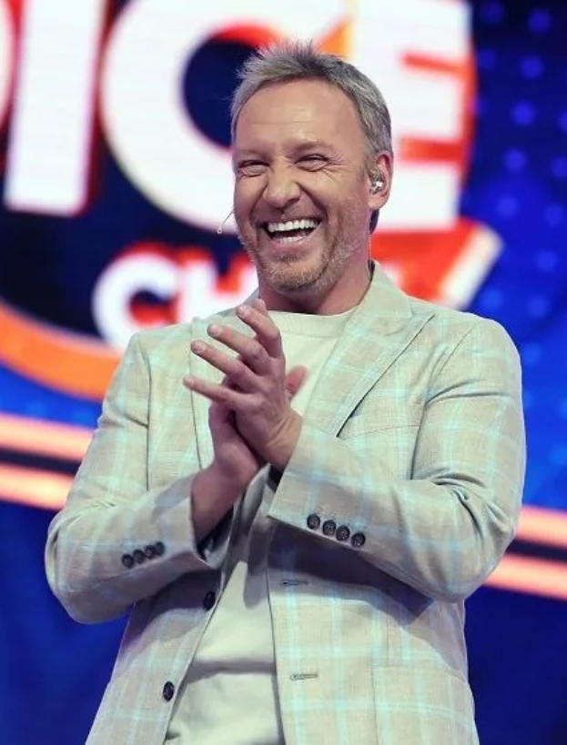 Ein Mann in einem Anzug, der auf einer Bühne klatscht, mit einem 'The Voice'-Schild im Hintergrund.