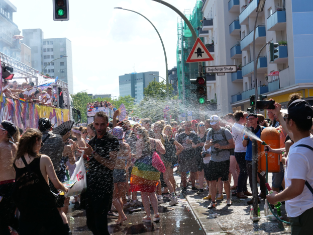Eine Gruppe von Menschen auf einer Straße bei einer Gay Pride Parade, einige spritzen sich gegenseitig Wasser ins Gesicht, während sie Gegenstände halten, mit einem Banner links und Gebäuden, Bäumen und Ampeln im Hintergrund.