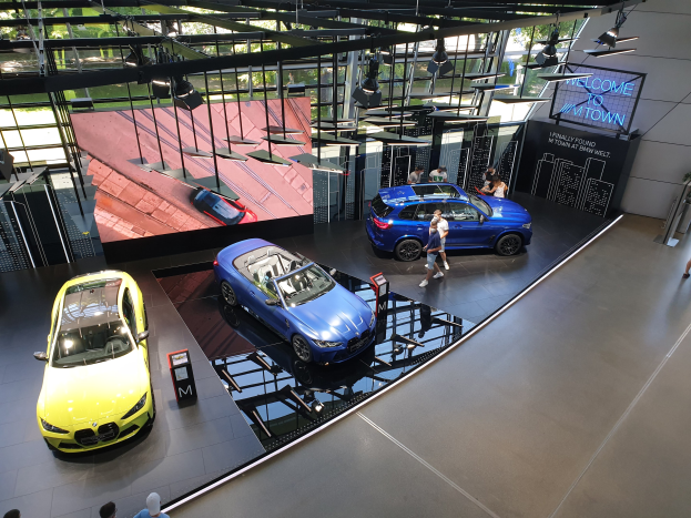 Eine Gruppe von Menschen im BMW Museum in München, Deutschland, mit Autos auf der linken Seite und einem Schild mit der Aufschrift "BMW Museum München" auf der rechten Seite, das auf eine Außenansicht von Bäumen, Gebäuden und Himmel durch Glasfenster blickt.