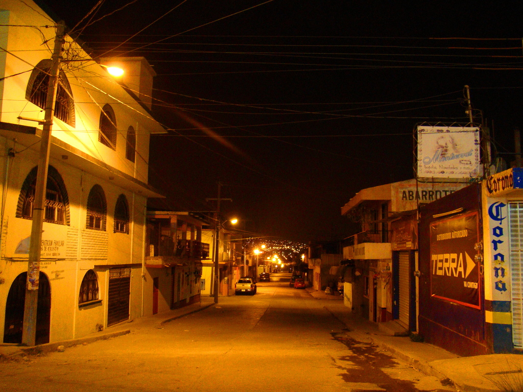 Eine Straßenszene mit Gebäuden und Strommasten auf beiden Seiten, einigen parkenden Fahrzeugen vor den Gebäuden und einer Straße in der Mitte.