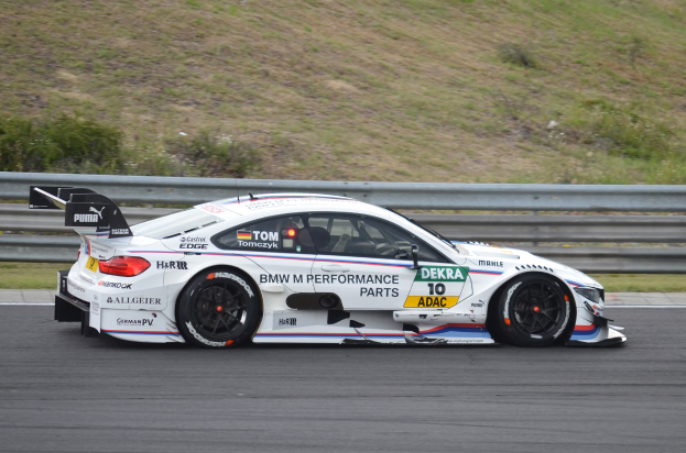 Ein BMW M4 DTM-Rennwagen fährt auf einer Rennstrecke, umgeben von grünem Gras, mit einer Begrenzung im Hintergrund.