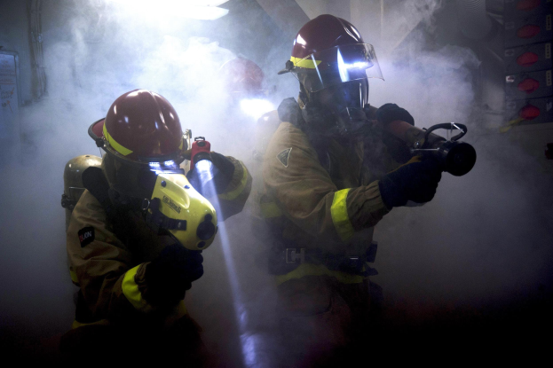 Zwei Feuerwehrleute in Schutzanzügen sprühen Wasser auf eine rauchgefüllte Wand, mit einer Tafel und verschiedenen Gegenständen in der Nähe.