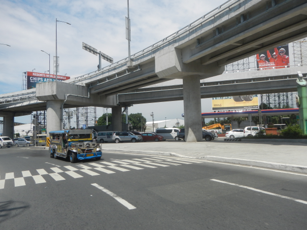 Eine vielbefahrene Hauptstraße mit Fahrzeugen, einer Zebrastreifen, einer Überführung mit Säulen, Laternen, Schildern mit Text und Bildern, Bäumen, Gebäuden und einem bewölkten Himmel im Hintergrund.