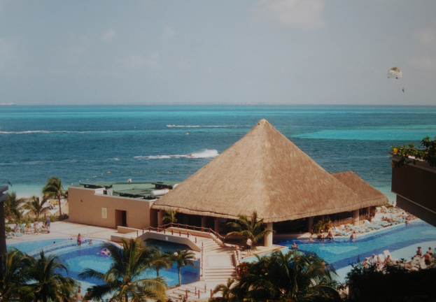 Ein Resort mit einem gro├čen Schwimmbad, umgeben von Palmen, Hütten, Treppen und Gel├Ąndern, mit Menschen im Pool und einem Ozean im Hintergrund mit einem bew├Âlktem Himmel und einem Fallschirm in der Luft.