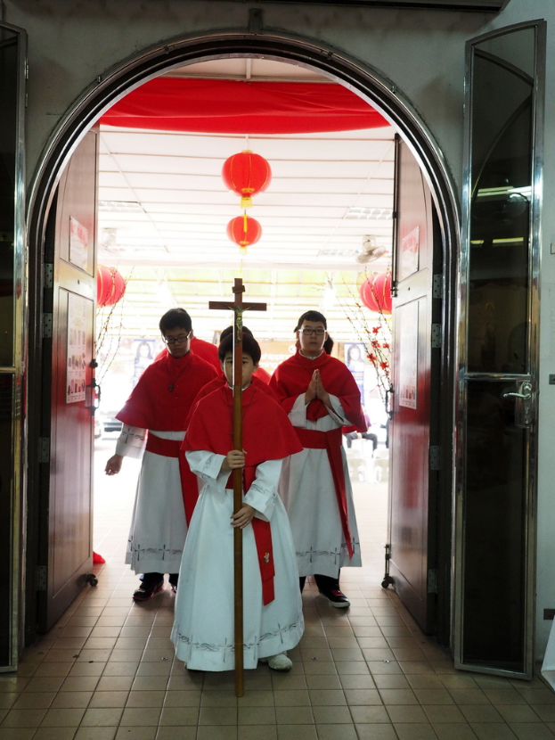 Menschen, die mit einem Kreuz in der Hand durch eine Kathedrale gehen, mit Türen links und rechts und Blumen und dekorativen Elementen im Hintergrund.
