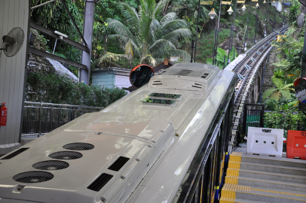 Ein Zug auf den Eisenbahnschienen mit Gebäuden, Polen, einem Ventilator, einem Feuerlöscher und einem Geländer auf der linken Seite, verschiedenen Gegenständen und einer Treppe auf der rechten Seite und Bäumen im Hintergrund.