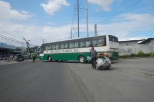 Grüner und weißer Bus fährt auf einer Straße mit Passanten, einem Radfahrer, Gebäuden, Bäumen und Strommasten im Hintergrund.