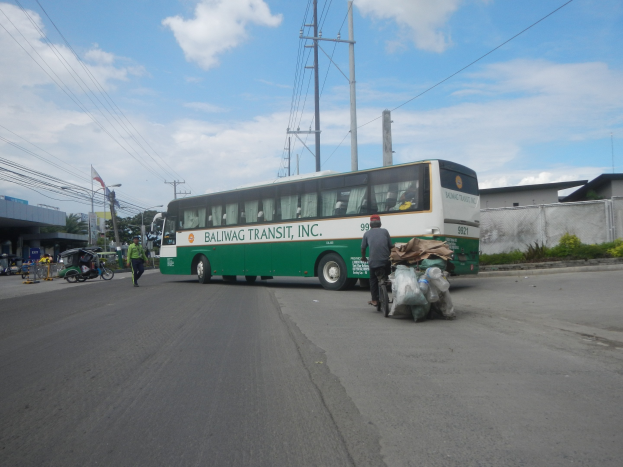 Grüner und weißer Bus fährt auf einer Straße mit Passanten, einem Radfahrer, Gebäuden, Bäumen und Strommasten im Hintergrund.