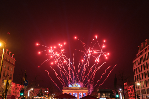 Eine Straße in Berlin an Silvester, voller Gebäude, Bäume, Laternenmäste, Ampeln, Schilder, Zelte und Menschen, mit einem Feuerwerk-himmel im Hintergrund.