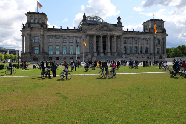 Gruppe von Menschen, die vor dem Reichstaggebäude in Berlin, Deutschland, Fahrräder fahren, mit Bäumen, Pflanzen und einem bewölkten Himmel.
