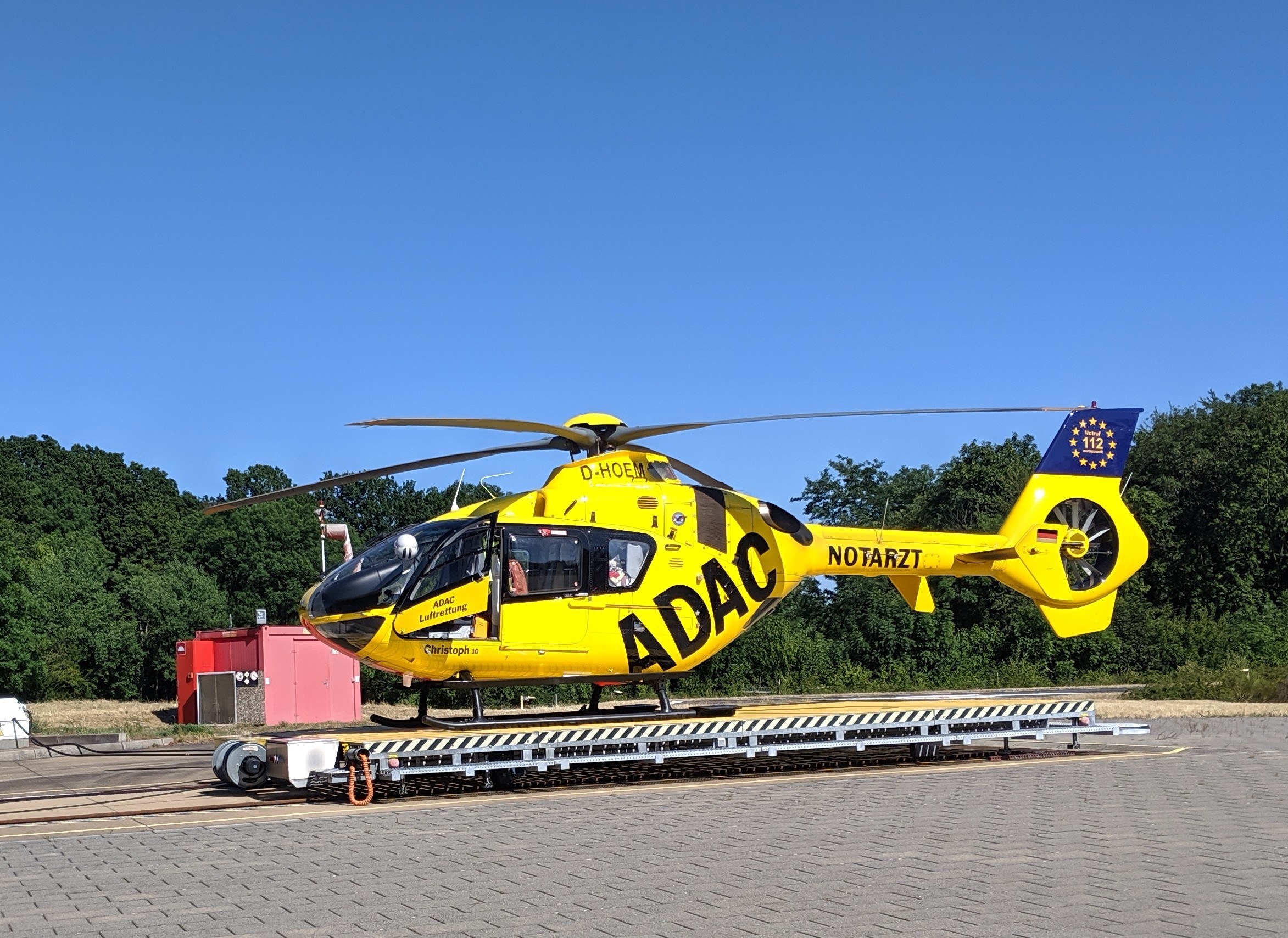 Ein gelber Hubschrauber sitzt auf einem Flughafen-Asphalt, umgeben von Bäumen und einem klaren blauen Himmel, mit verschiedenen Gegenständen, die mit dem Hubschrauber in Verbindung stehen.