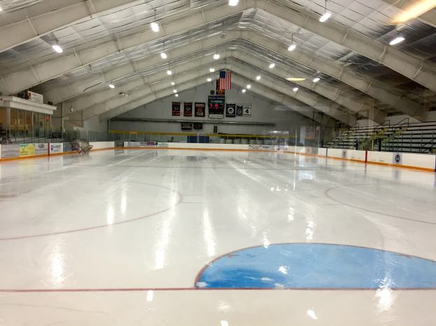 Große Indoor-Eisfläche mit Deckenbeleuchtung, Texttafeln, einer Wand, Treppen mit Geländern und einigen Menschen, was auf eine Sportanlage hinweist, die wahrscheinlich für Hockey verwendet wird.
