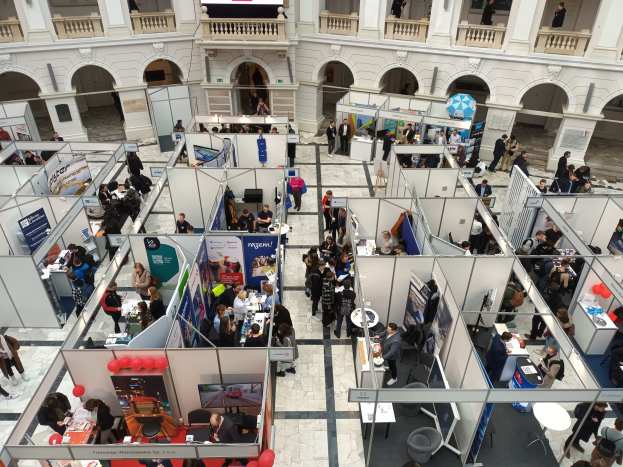 Eine große Gruppe von Menschen auf einer internationalen Messe mit Ständen, Bannern, Tischen, Stühlen, Ballons und einem Gebäude mit Bögen, Säulen, Geländern und einem Bildschirm im Hintergrund.