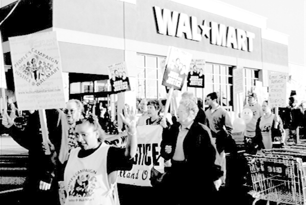 Ein Schwarz-Weiß-Bild von protestierenden Menschen, die Plakate halten, mit einem Einkaufszentrum im Hintergrund.