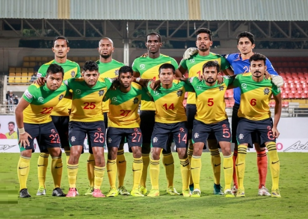 Eine Gruppe von Männern auf einem Fußballfeld, die einen Sieg feiern, umgeben von Werbetafeln, Stühlen, Geländern, Pfosten, Bäumen und einer Hütte im Hintergrund.