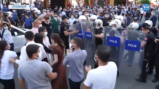 Eine Gruppe von Menschen steht einer Reihe von Polizisten in Helmen und Schilden gegenüber, mit einem Auto links, einer Person mit einer Kamera rechts und Gebäuden, Bäumen und Texttafeln im Hintergrund in Ankara, Türkei.