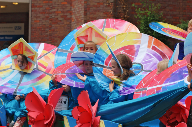 Eine Gruppe von Kindern in bunten Kostümen fährt auf einem Umzugswagen, hält Schirme, mit einem Gebäude, Pflanzen und Lichtern im Hintergrund.
