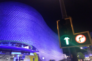 Das Selfridges Einkaufszentrum in Birmingham, England, ein Gebäude mit Säulen und Lichtern, umgeben von Bäumen, Laternen und Fahrzeugen auf der Straße, mit einer Ampel vorne und dem Himmel im Hintergrund.