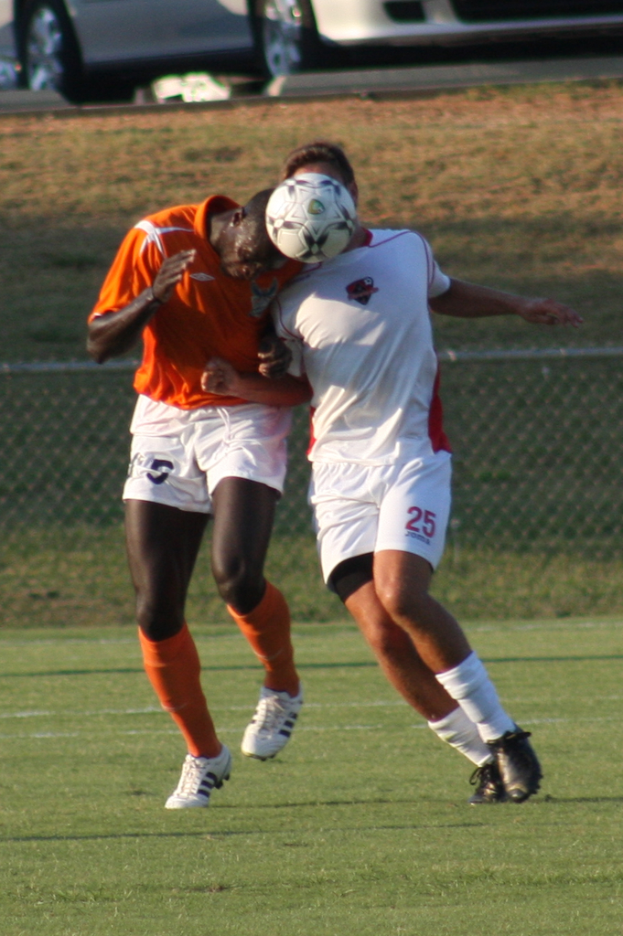 Zwei Spieler auf einem umzäunten Platz, einer im orangen Trikot schießt den Ball mit dem Kopf, mit Autos und Gras im Hintergrund.
