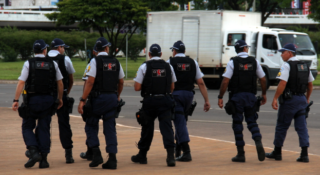 Eine Gruppe von Beamten in Uniform, einschließlich Mützen, Hemden, Schutzwesten, Uhren, Hosen und Schuhen, geht auf einer Straße mit einem geparkten Lkw auf der rechten Seite neben einem Zaun. Im Hintergrund sind Gebäude, Bäume, Pflanzen, Pfosten, Banner und eine Brücke im oberen rechten Eck zu sehen.