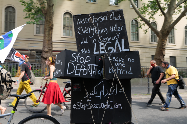Eine Gruppe von Menschen geht mit Fahrrädern die Straße entlang, einige tragen Taschen und einer hält eine Fahne, im Hintergrund sind Bäume, ein Gebäude mit Fenstern und ein Schild mit der Aufschrift "Stoppt Abschiebung".