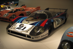 Ein Porsche 917K Le Mans Rennwagen wird in einem Museum ausgestellt, umgeben von anderen Sportwagen und Menschen, mit einer Wand im Hintergrund und einem Papier mit Text auf der linken Seite.
