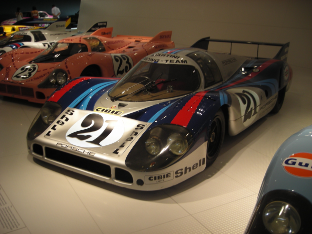 Ein Porsche 917K Le Mans Rennwagen wird in einem Museum ausgestellt, umgeben von anderen Sportwagen und Menschen, mit einer Wand im Hintergrund und einem Papier mit Text auf der linken Seite.