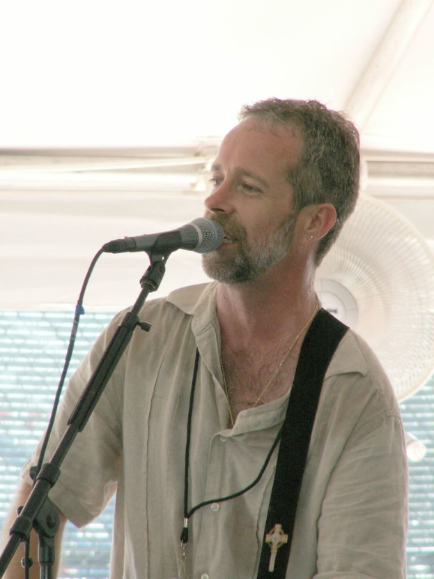 Ein Mann singt in ein Mikrofon mit einem Tischventilator im Hintergrund.
