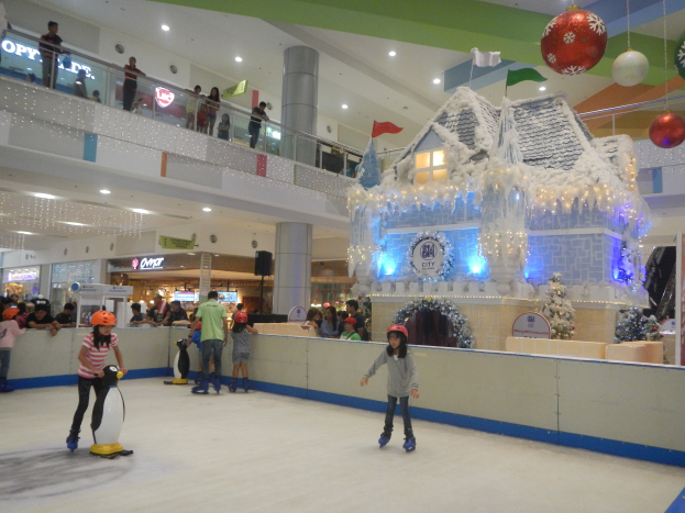 Menschen, die auf einer Eisbahn in einem Einkaufszentrum Schlittschuhlaufen, umgeben von festlichen Dekorationen, während andere auf Geländern und Säulen stehen.
