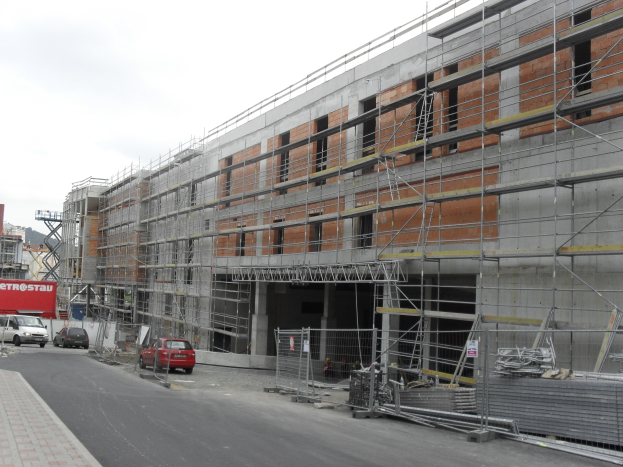 Großes Gebäude im Bau mit Gerüst, umgeben von Fahrzeugen auf der Straße, einem Fußweg auf der linken Seite und einem klaren blauen Himmel im Hintergrund.