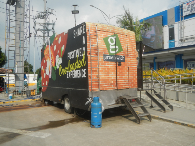 Lkw vor einem Gebäude mit einer Tafel, auf der "Teilen Sie ein positiv überladenes Erlebnis" steht, mit Stufen, einem Zylinder auf dem Boden, Geländern, Strommasten mit Drähten, Laternen, Bäumen und einem bewölkten Himmel.