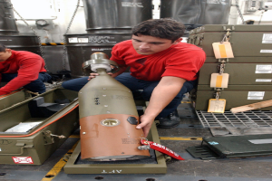 Zwei Männer in roten Hemden arbeiten an einer großen Metallfragmentierungsbombe, mit Kisten, Ketten und anderen Gegenständen im Hintergrund.